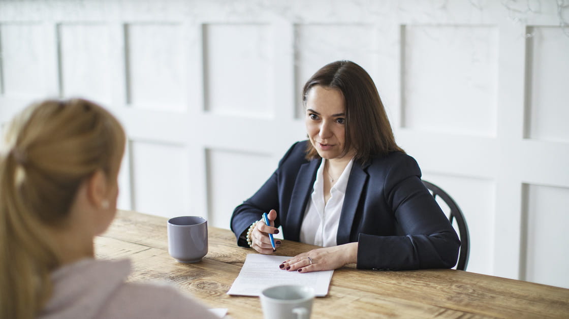 A white woman with brown hair is sitting at a table. She is wearing a navy blue jacket and a white shirt. She is holding a paper and pen and talking to someone in across the table. 