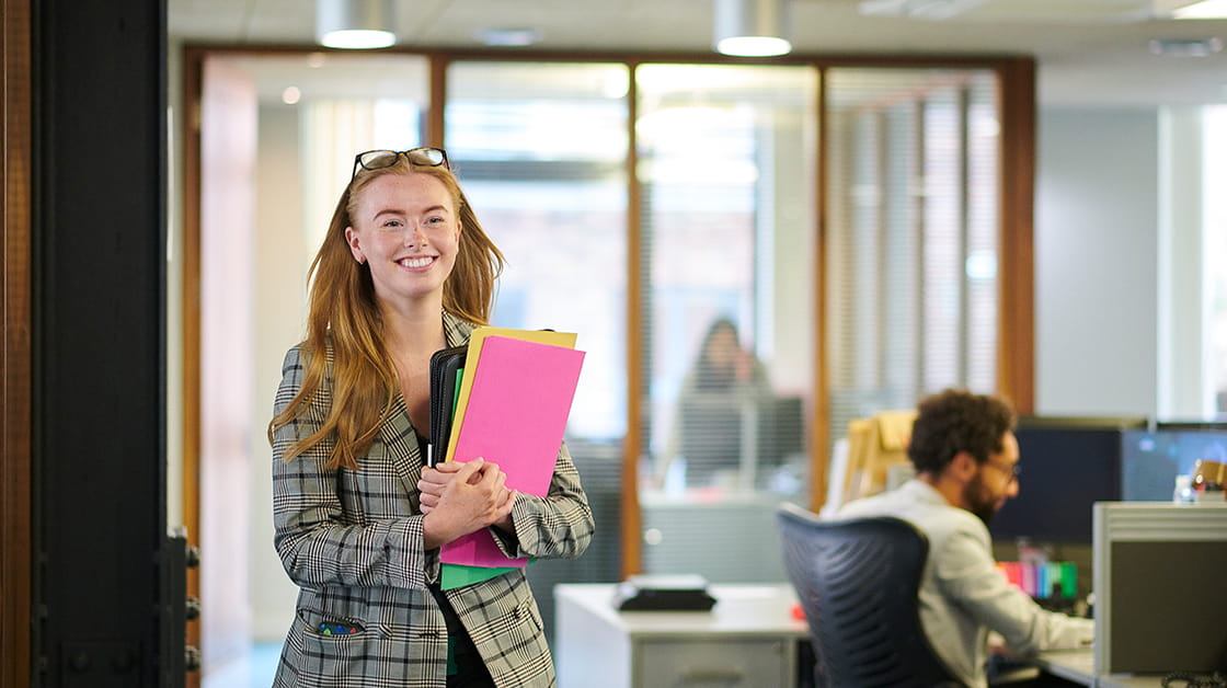 A smiling paralegal walks through an office holding paperwork.