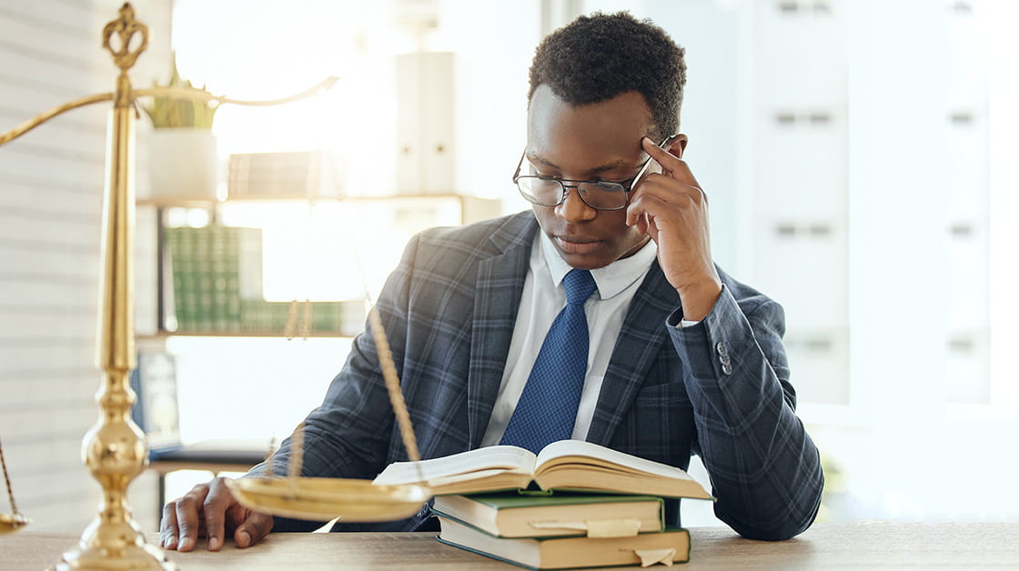 Young black male lawyer reading from a pile of books with post-its marking places in brightly lit office. He is wearing glasses and a checked suit with blue tie. Next to him is a set of scales.