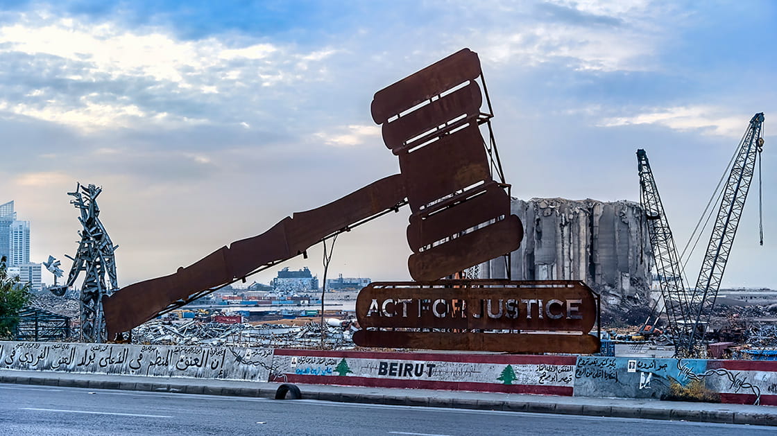 "Act for Justice" gavel sculpture among memorials at Beirut Port Explosion site, Lebanon (December 2021). In the background, there are cranes and wreckage of port buildings.