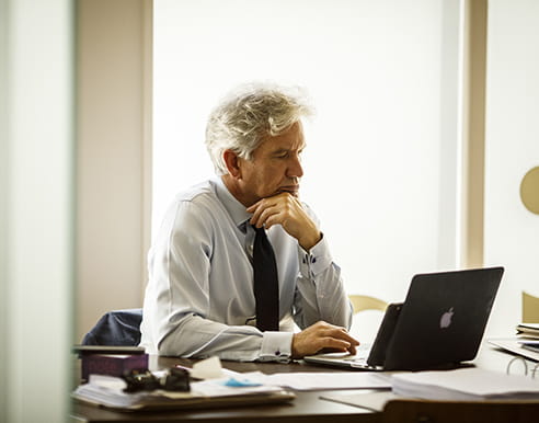 A man with grey hair, wearing a white shirt and blue tie, is sitting at a desk with his hand resting on his chin, looking at a laptop