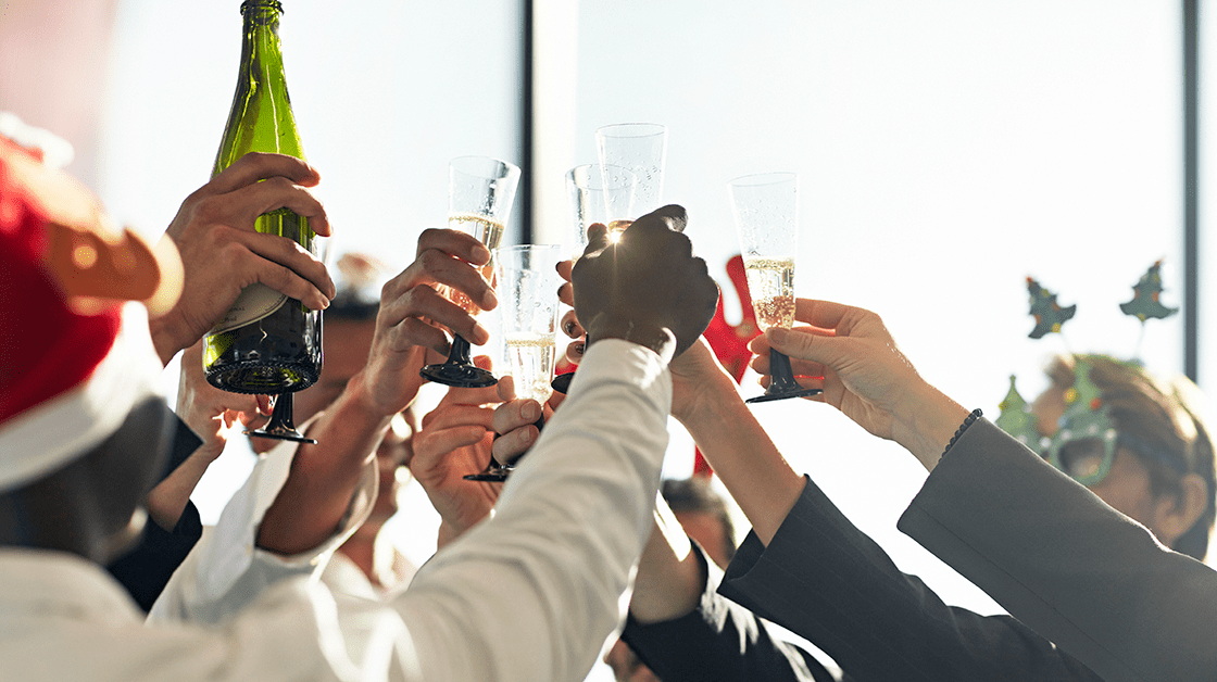 A group of colleagues celebrate at the office Christmas party wearing festive hats and raising glasses of champagne.