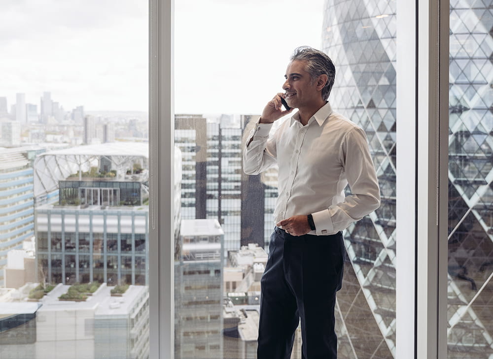 Grey-haired lawyer chats on phone with City of London skyline view behind him.