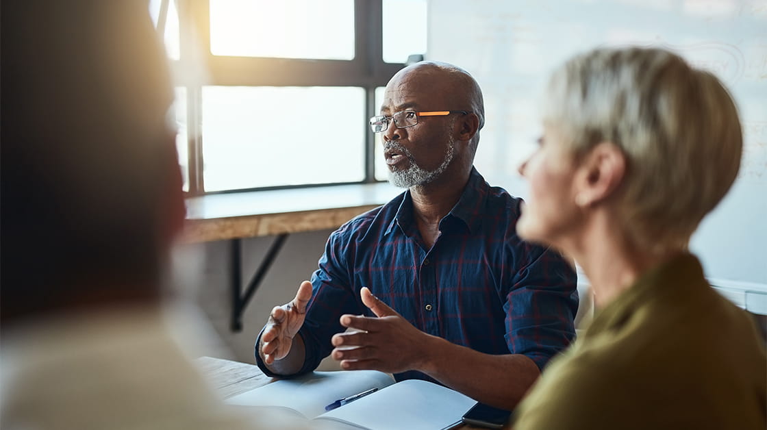 A group of colleagues sit in a meeting room discussing their ideas. The speaker is a Black man with cropped grey hair, glasses and a beard, who wears a dark blue shirt. In the foreground, a white woman with short grey hair wearing a green shirt listens intently.