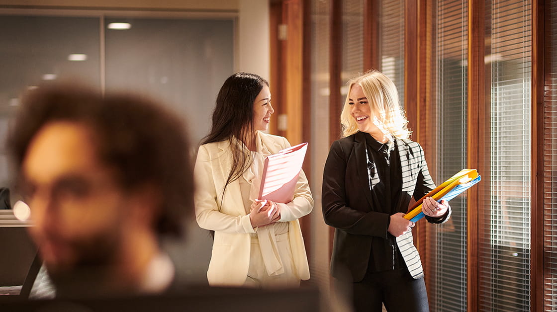 Two colleagues walk together along a corridor, carrying files and talking.