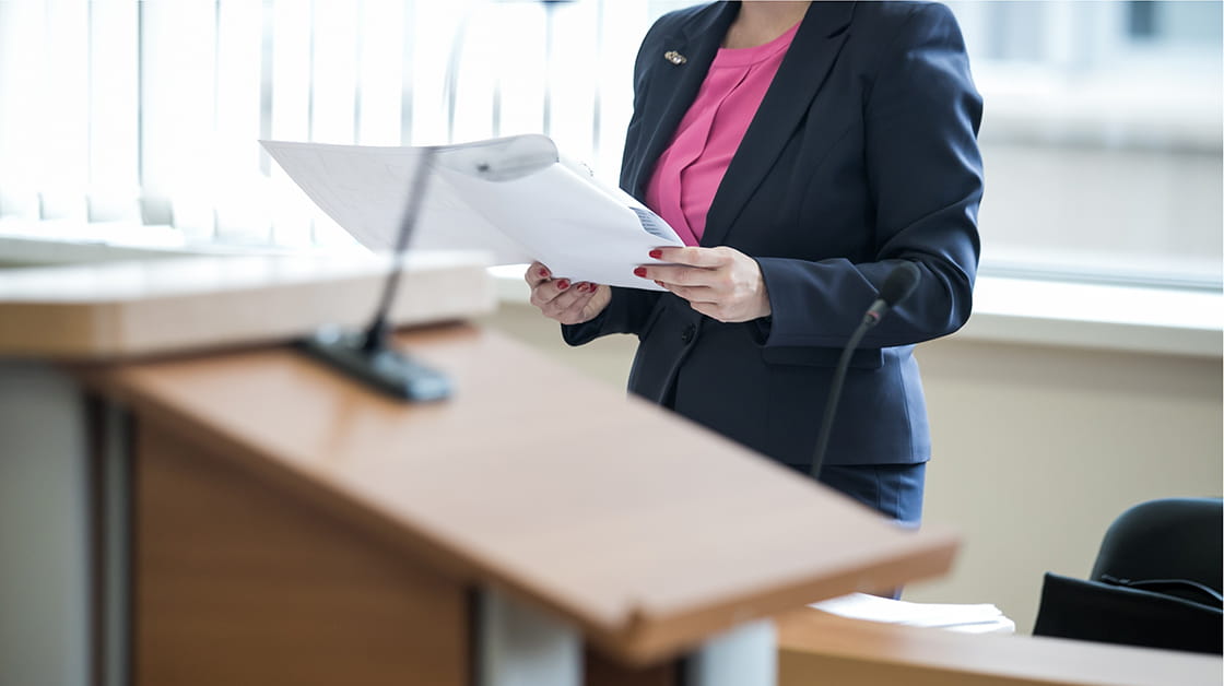 Lawyer in court holding papers while cross-examining witness. She is wearing a pink top and dark suit, visible from the shoulders down