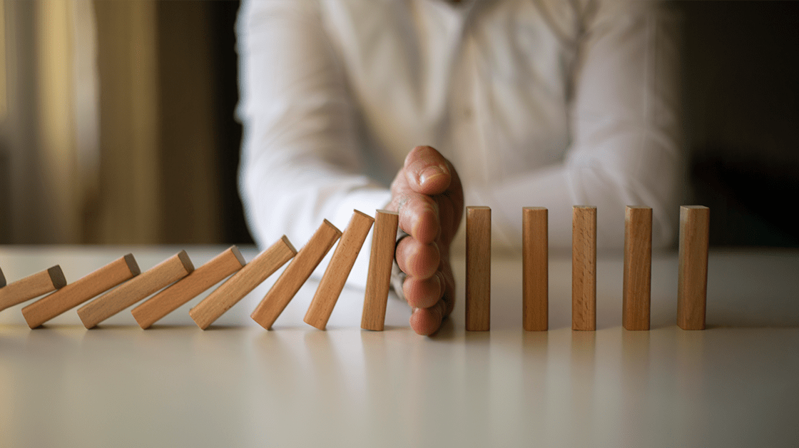 A chain of falling wooden domino blocks is prevented from falling further by the hand of a black man wearing a white shirt.