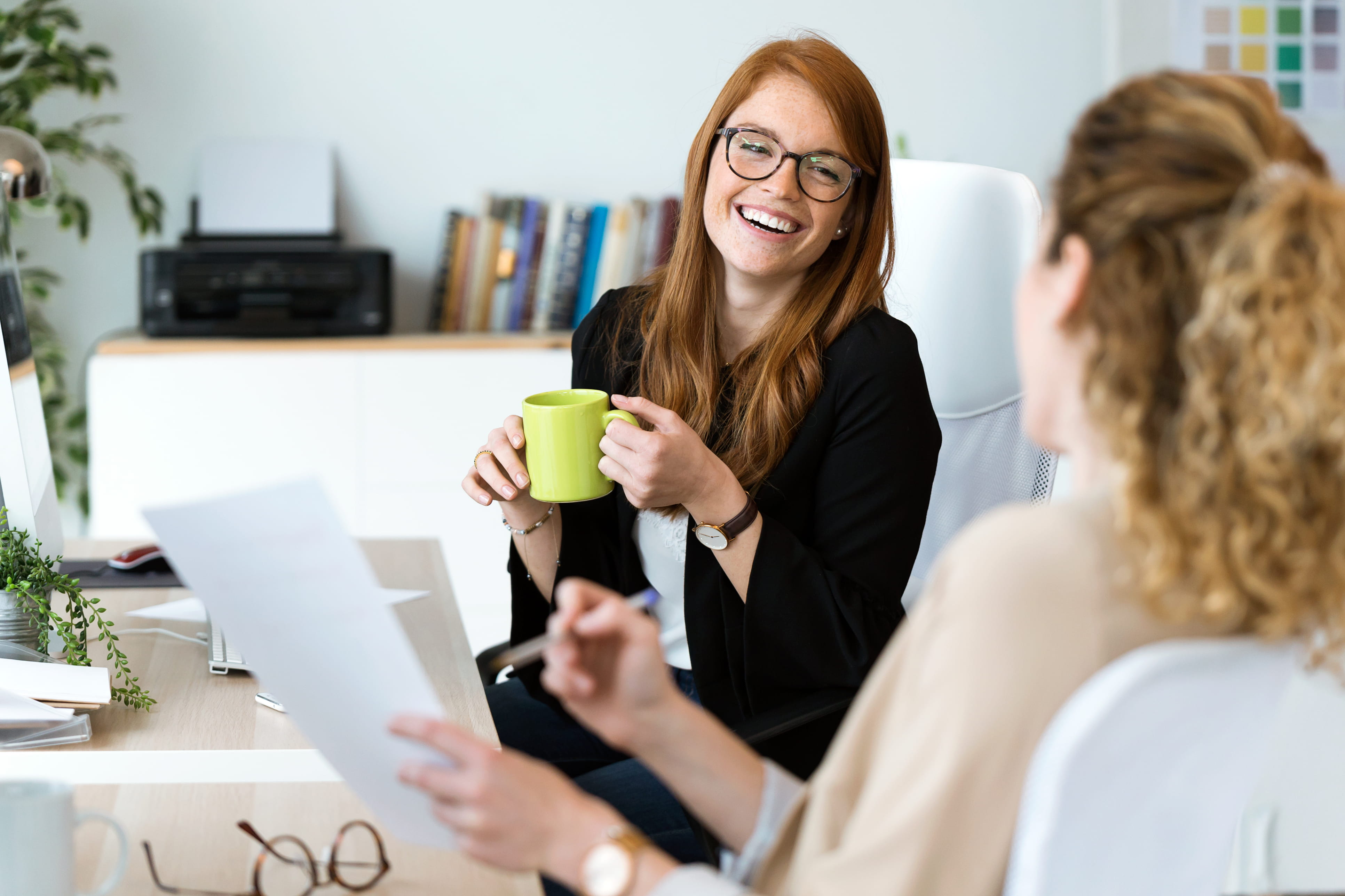 A white woman with ginger hair and glasses is sitting in an office. She is smiling and holding a green mug with two hands. She is wearing a black jacket and white blouse. She is talking to her colleague who has blonde, curly hair. 