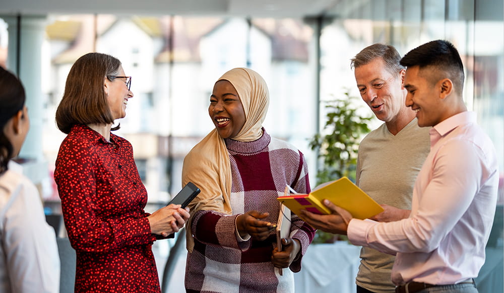 A mixed ethnic group of business professionals at a networking conference/meeting room in the North East of England. They are standing holding books/digital tablets smiling while discussing business together.