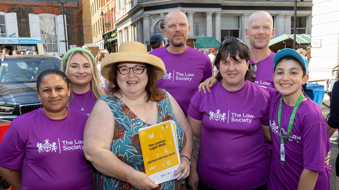 A diverse group of people, wearing purple t-shirts branded with European Union logo are standing outside in a group and smiling. In front of them is a white woman in a blue, patterned top. She is wearing glasses and a sun hat. 