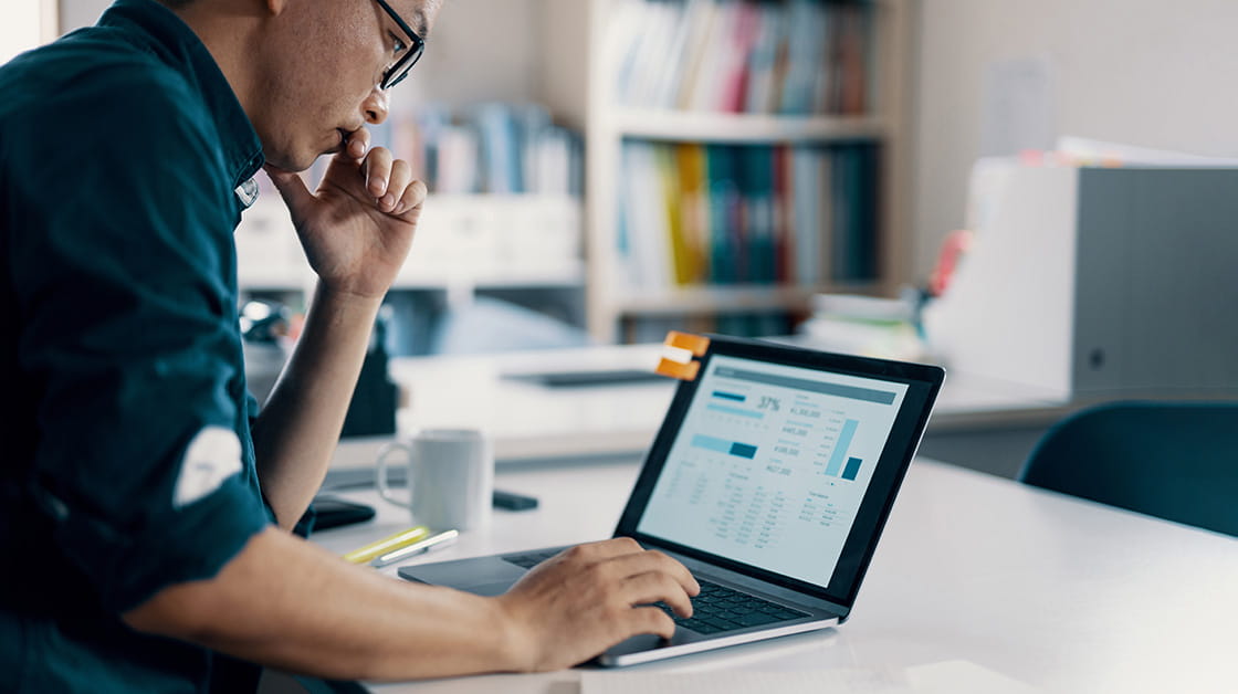 A man of East Asian heritage with short black hair sits at a laptop looking at financial projects. He wears glasses and a dark blue shirt.