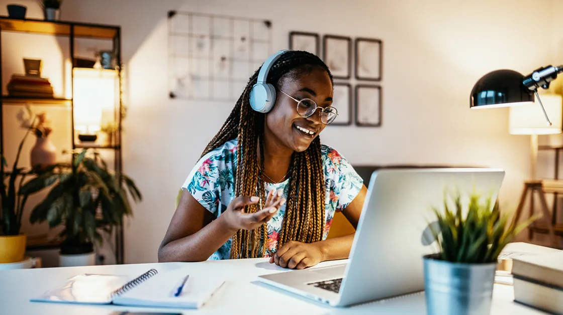 A black woman with long brown, braided hair is sitting at her desk at home. She is wearing glasses and big headphones and smiling at her laptop. There is a small plant on her desk and a plant by some shelves behind her.