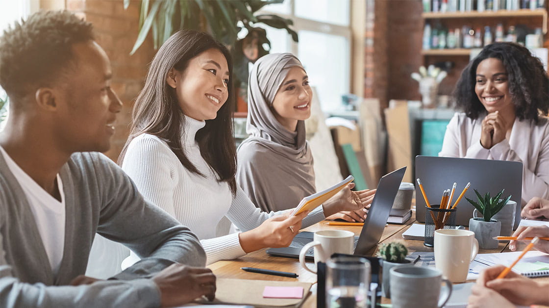 Group of diverse young professionals sat around table talking and smiling