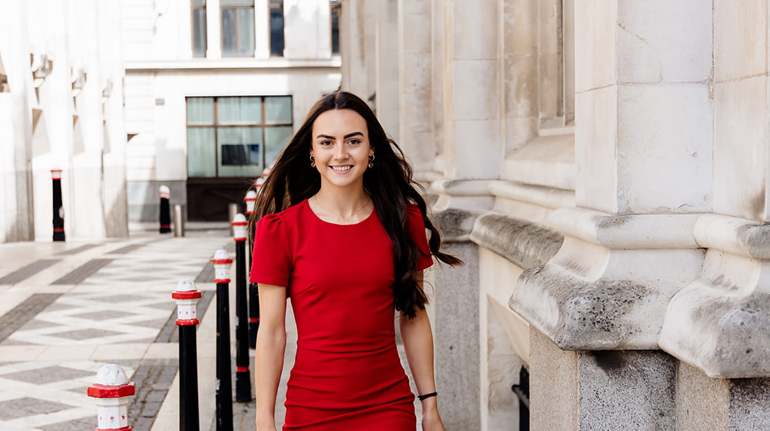 Holly Moore is a white woman with long, wavy, dark brown hair. She is smiling widely and walking down a cobbled street, surrounded by stone buildings. She wears a red dress.