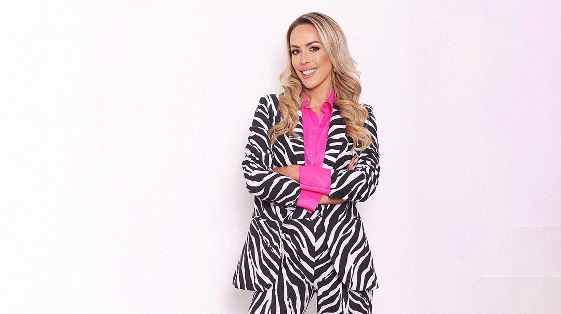 Jodie is a white woman with blonde hair. She is wearing a zebra patterned suit and pink shirt. She is standing with her arms crossed in front of a white background. 