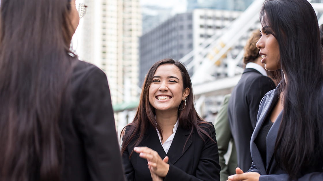 Young asian woman laughing and clapping hands together while speaking to two colleagues. They all wear dark suits and are standing outdoors with high-rise City buildings in the background.