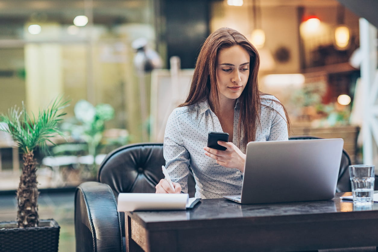 A woman sits at a desk with a closed mouth smile. She is looking at a laptop, holding a phone and writing on a piece of paper. 