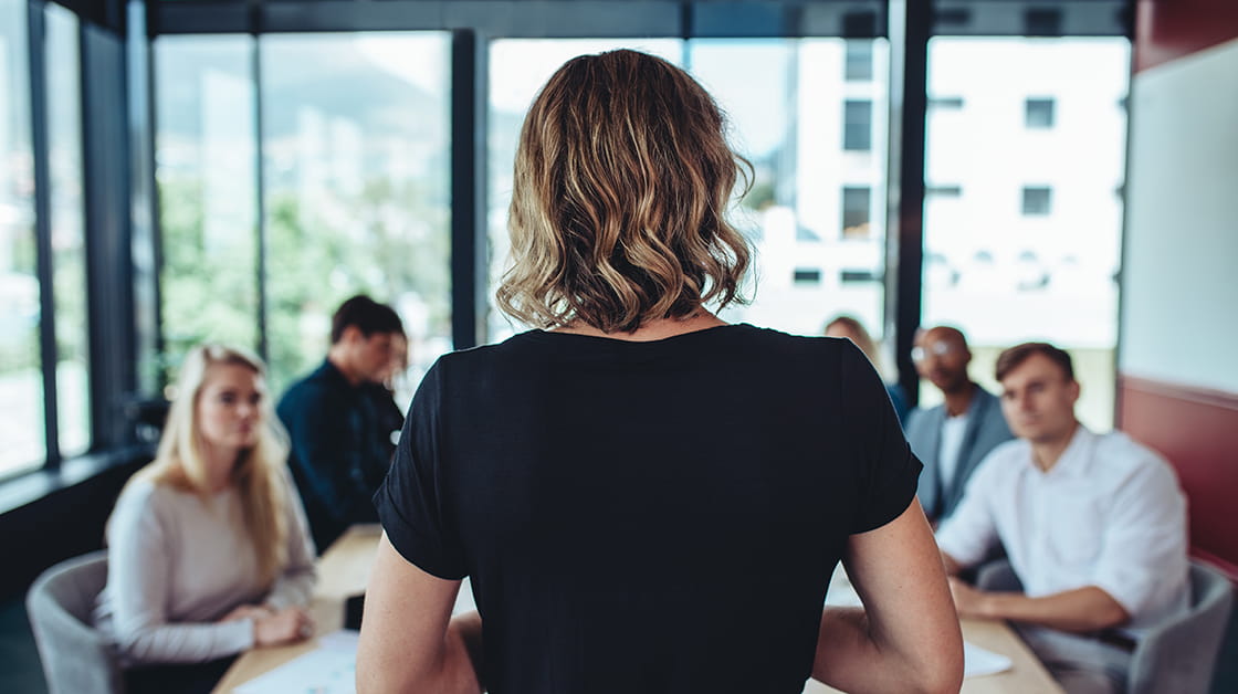 A new business owner stands at the head of a meeting table with her back turned as she speaks with colleagues. She is a white woman with short, curly, dark blonde hair. She wears a black shirt and grey suit trousers.