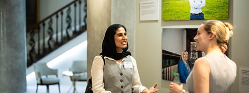 A woman in a black head scarf, wearing a white shirt and grey button down sweater is talking to a white woman with blonde hair