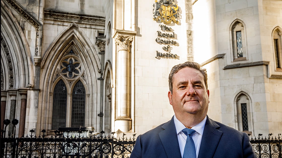 Nick Emmerson standing in front of the Royal Courts of Justice, London on a bright sunny day. Nick is a white man with dark hair, wearing a blue suit and tie. 