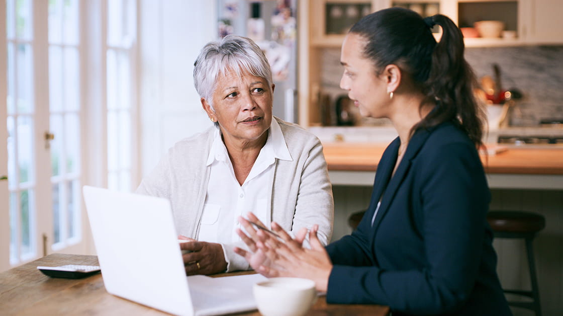 An older mixed race woman with short, grey hair wearing a white shirt and beige cardigan sits at a desk receiving advice. She speaks to her financial adviser, a woman with long dark brown hair in a slicked-back ponytail. She wears a dark suit and is explaining a concept by gesturing.