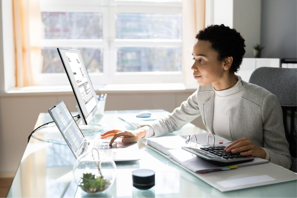 A black woman with short black hair is wearing a grey jacket and white top. She is working in front of two computer screens. 