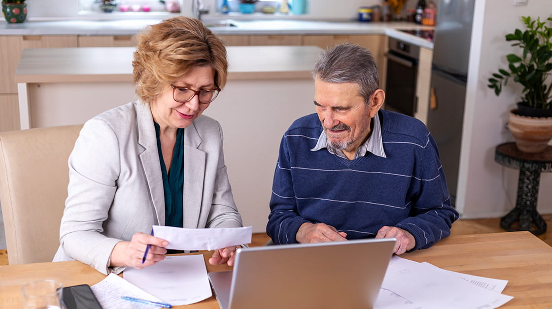 Mature woman helping senior man with paperwork in his kitchen. She is a white woman wearing glasses and a pale suit, he is a white older man with a grey moustache and a blue jumper over a shirt