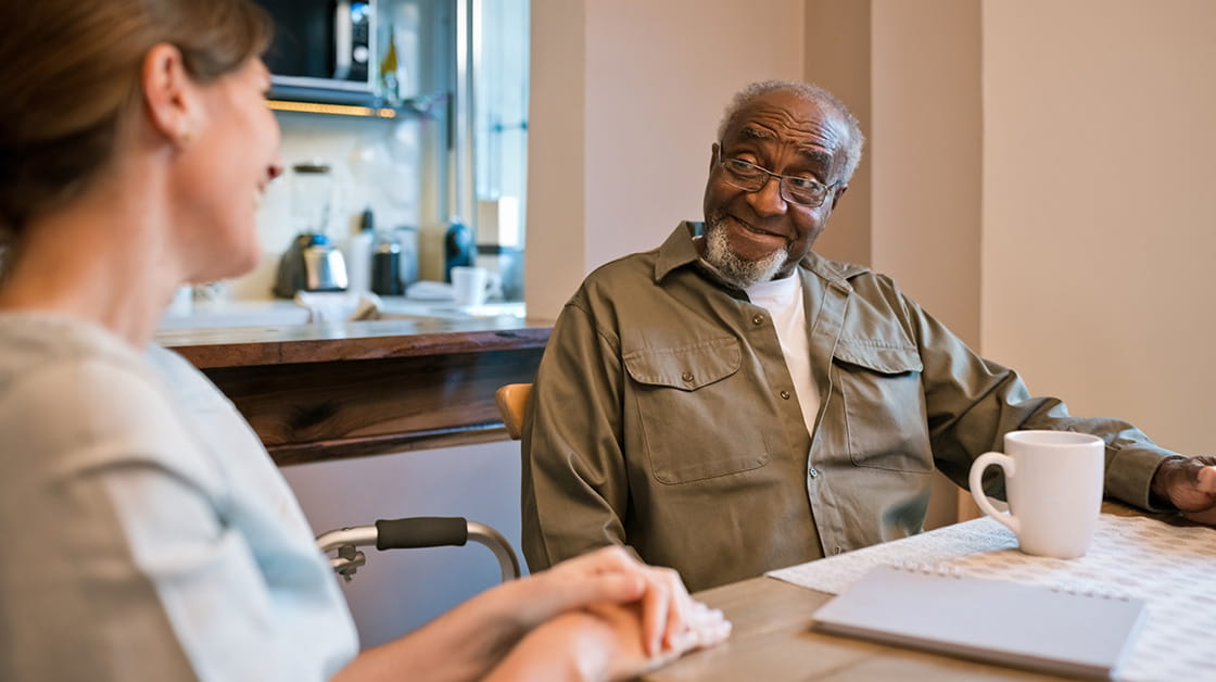 An elderly male client sits next to a professional woman. He is a Black man with balding grey hair and a beard. He wears glasses and a khaki shirt with a white t-shirt. She is a white woman with dark brown hair. She wears a pale blue shirt.