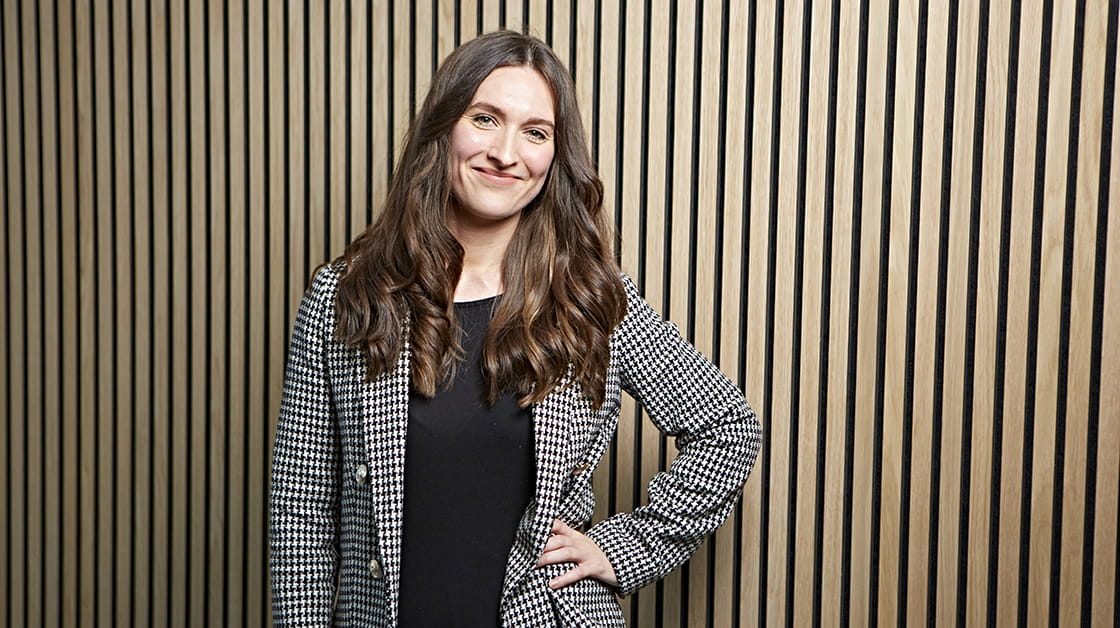 Rebekah Sutcliffe is a white woman with long, wavy brown hair. She is smiling and wears a houndstooth suit over a black blouse.