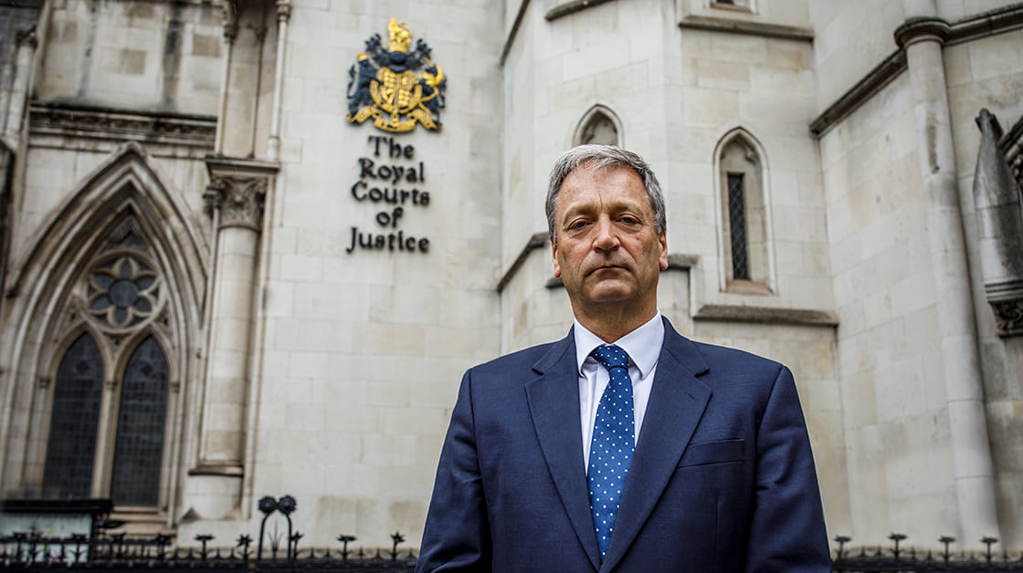 Richard Atkinson stands in front of the Royal Courts of Justice, London. Richard is a white man with short dark grey hair. He has a grave expression and wears a navy suit, white shirt and navy polka dot tie.