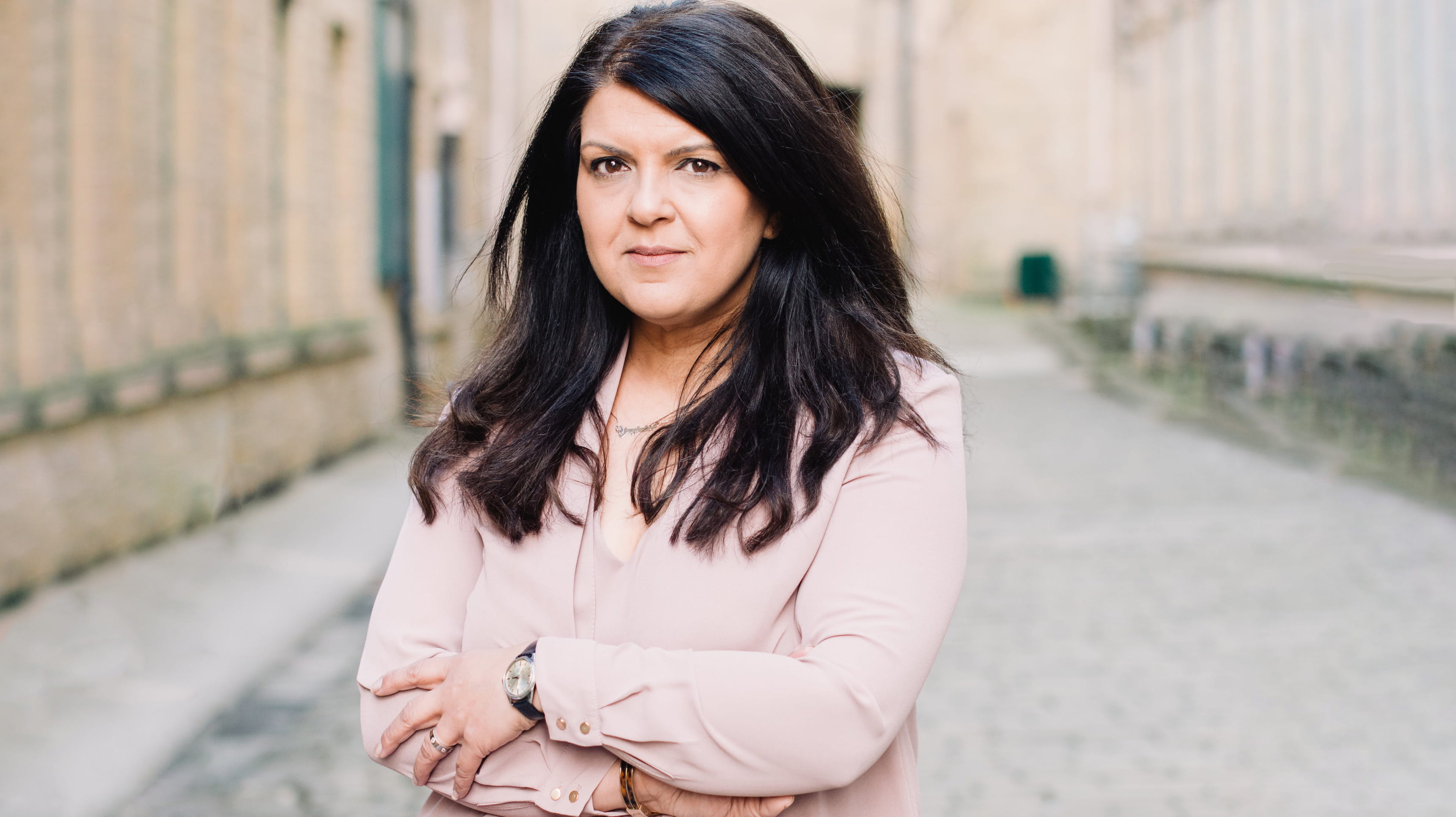 Sarah is a South Asian woman with dark brown hair. She is wearing a light pink blouse and a watch with a black strap. She is standing outside, smiling closed mouthed with her arms folded.