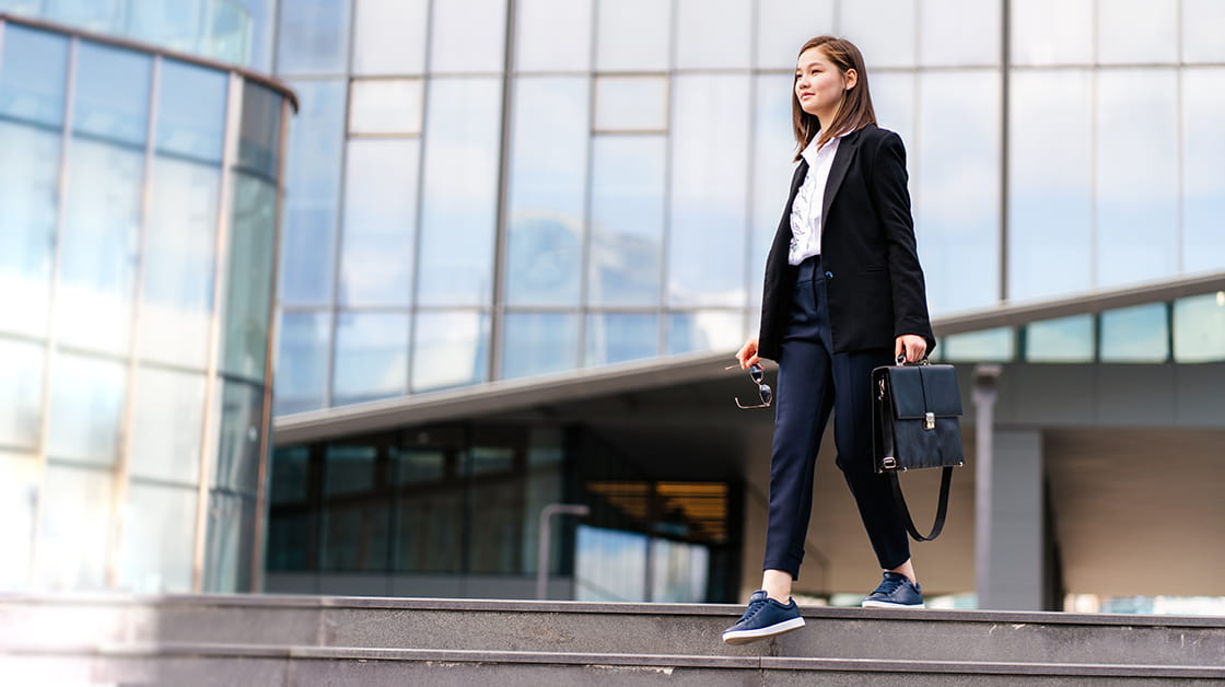 Young asian woman in smart casual business wear walking down steps outside office block - she's holding a briefcase, looking into the distance and smiling