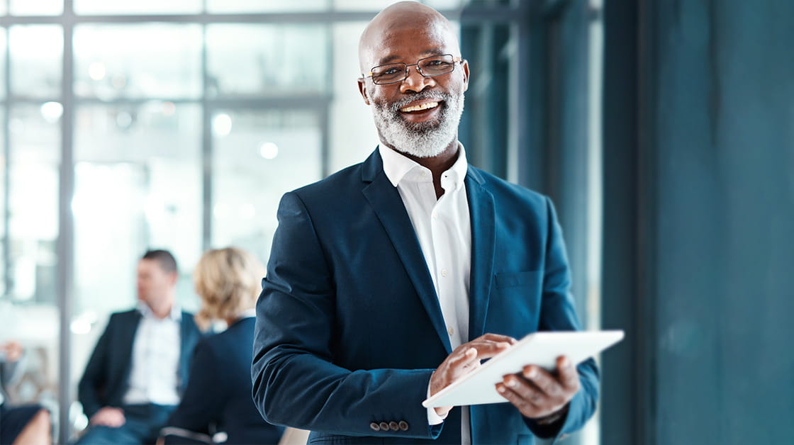 An older Black man with a shaved head and short grey beard stands in front of colleagues in a meeting room. He is smiling widely and wears a navy suit and white shirt. He holds a tablet.