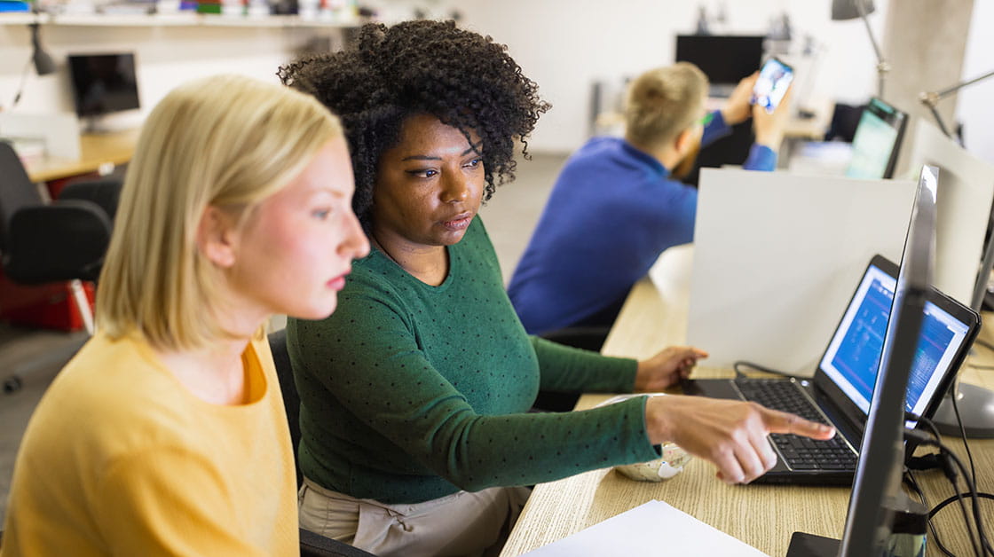 Two colleagues sit at a desk looking at a computer screen: a white woman with short blonde hair wearing a yellow shirt and a black woman with short afro hair wearing a green shirt.