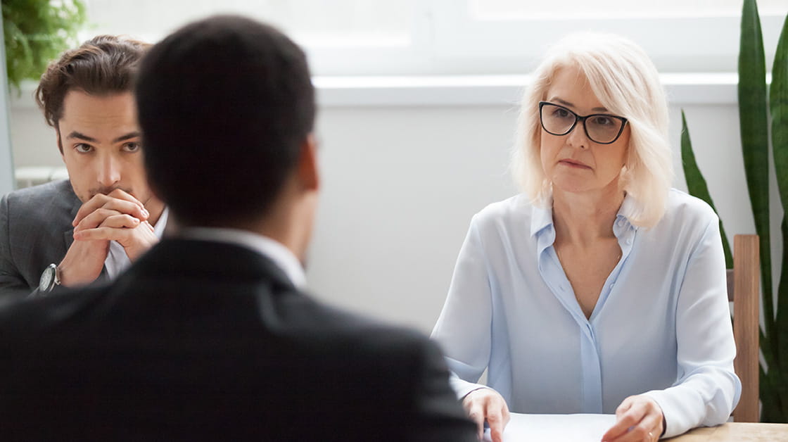 Serious meeting / interview between three professionals: white woman with platinum blonde hair and thick-framed glasses looks intently at man in suit across table. Next to her, younger white male colleague looks at interview over interlocked fingers.