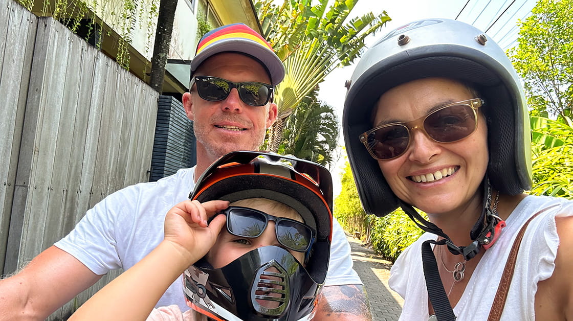 Simone Ritchie and her husband and son, wearing scooter helmets and sunglasses, with a tropical greenery behind them on holiday in Bali