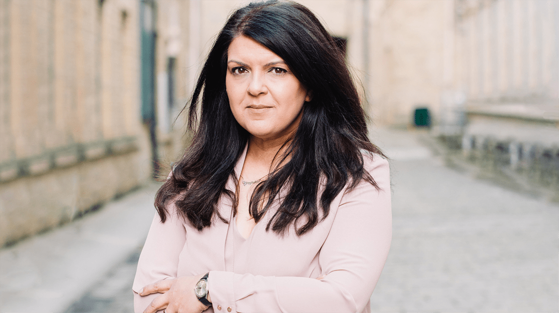 Sarah is a south Asian woman. She is wearing a pink suit and a black watch, she has her arms crossed. She has a neutral expression.