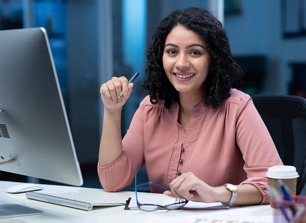 Woman working at her desk - she has dark curly hair and is wearing a dusty pink blouse