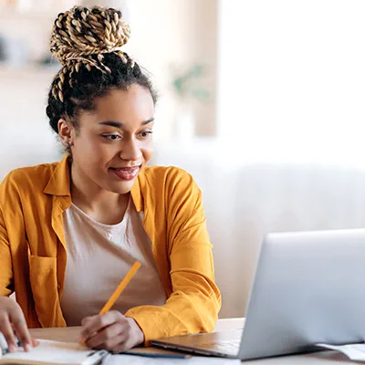 A smiling young mixed race woman with braided hair is studying using a laptop and notebook