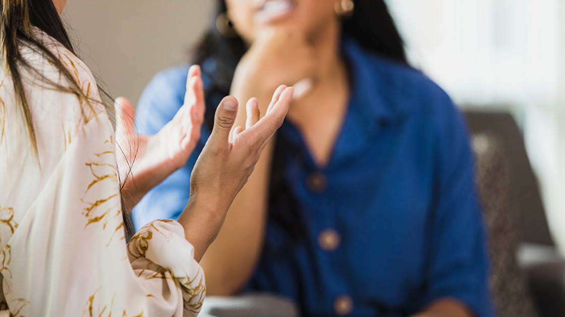 A female client sits opposite a woman professional. They are both of South Asian heritage and have long, straight black hair. The client wears a white and gold patterned blouse and the professional wears a navy shirt.