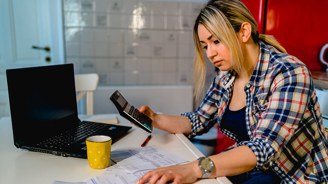 Young woman sitting in kitchen, staring at calculator next to open laptop