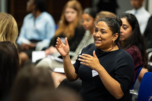 A black woman is standing in a room with a diverse group of people, she is wearing a black dress, speaking and gesticulating with her hands. 