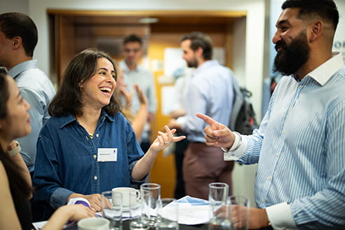 A white woman with short brown hair, wearing a denim shirt, is talking to an Asian mean, wearing a blue and white shirt. They're standing by a table and gesticulating with their hands.