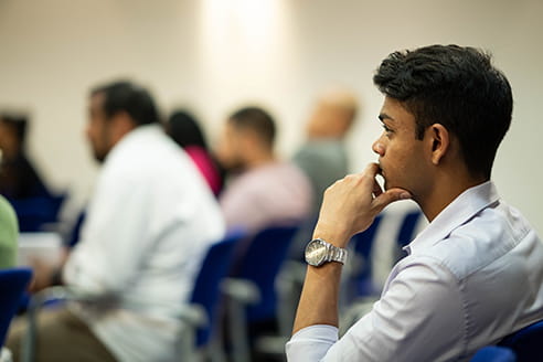 The side profile of a man with short brown hair, wearing a light blue shirt and a watch, holding his hand up to his mouth.