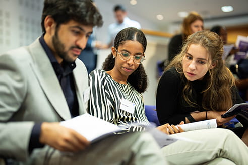 Three people are sitting, looking at a piece of paper. On the left, an Asian man with a beard is holding the paper, in the middle, a black woman wearing glasses and a stripy dress is looking at it and on the the right a white woman with long brown hair is leaning over and looking at the paper.