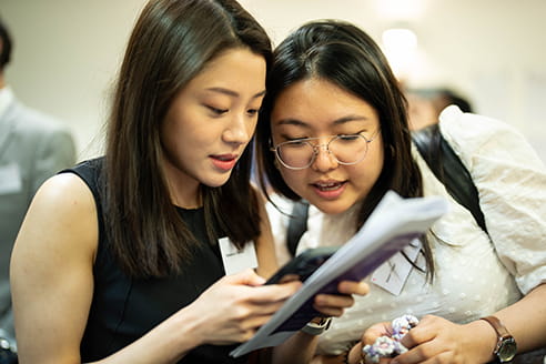 Two Asian women are standing together, looking at a phone. The woman on the left has long black hair and is holding the phone, the woman next to her has short black hair and glasses.