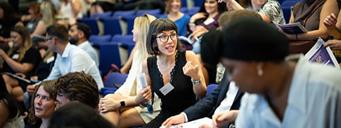 A diverse group of young professionals in a lecture theatre, a woman with short black hair and glasses is speaking and pointing. 