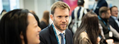 A white man with blonde hair and a beard, wearing a black suit, white shirt and blue tie