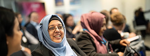 A woman in a blue head scarf and glasses is sitting down and smiling