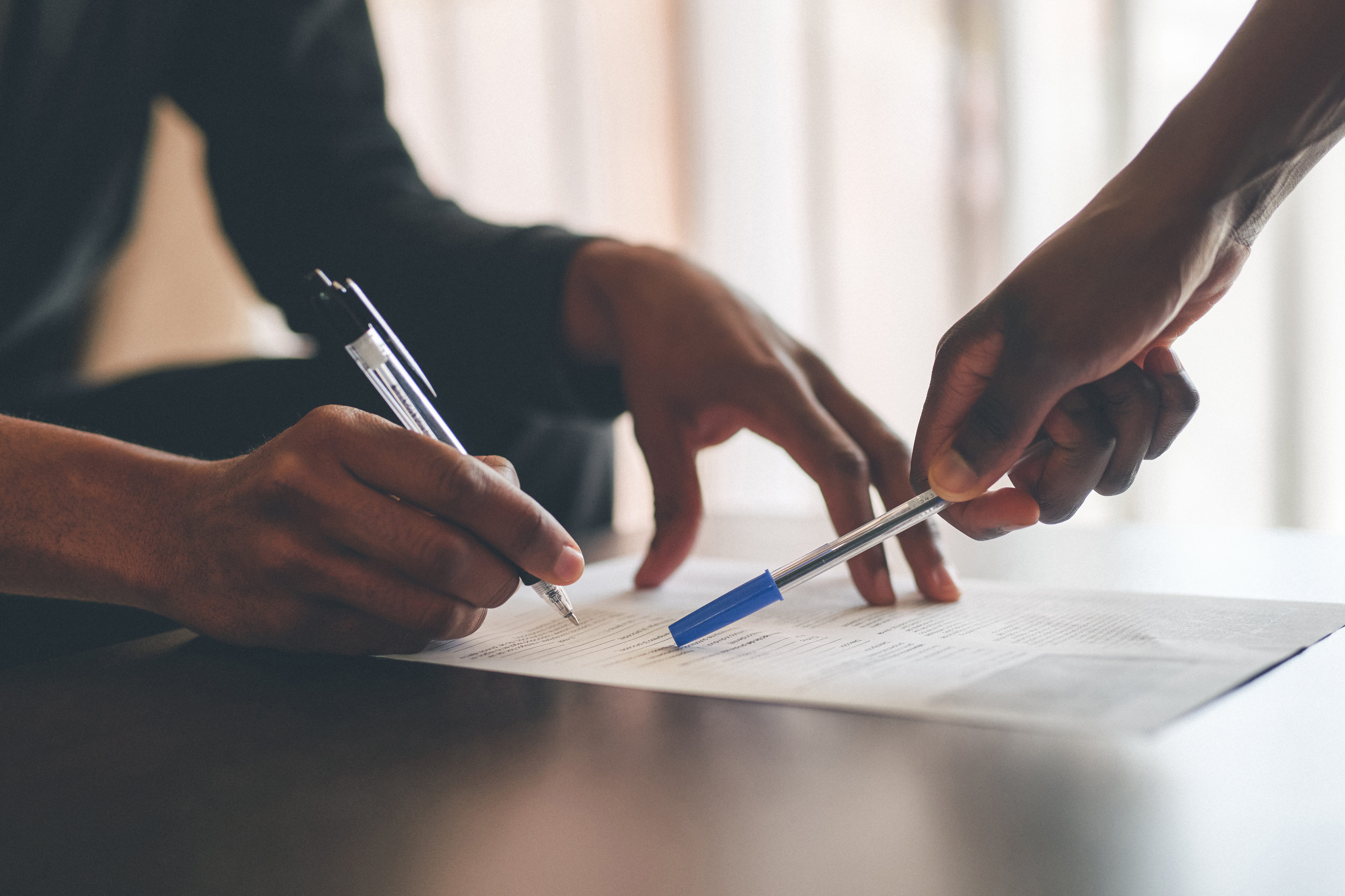 An image of a sheet of paper on a shiny table surface. There are words on the page, and two people are pointing to areas of the page with pens. You cannot see their faces, only their hands.